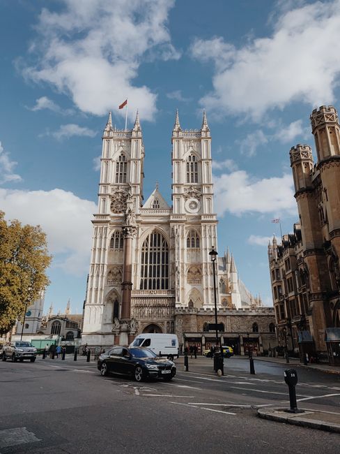 The image shows the facade of Westminster Abbey, a historic Gothic cathedral in Westminster, London, with its tall stone towers, intricate architectural details, large stained glass windows, and a central entrance arch. The sky is partly cloudy with patches of blue, and the surrounding area includes a busy street with moving cars, a white van parked near the cathedral, and pedestrians walking along the pavement. To the right, there are older brick buildings with decorative turreted rooftops. In the foreground, there are black metal bollards and streetlights along the roadside. The scene captures a typical daytime view of a prominent London landmark with urban traffic and pedestrians, while indicating a context where house removals or moving services, like those offered by Westminster Man and Van, might be relevant amid the city's busy environment.