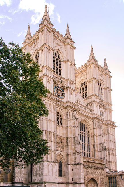 The image shows the facade of Westminster Abbey, a historic Gothic cathedral in Westminster, London, with its tall stone towers, intricate architectural details, large stained glass windows, and a central entrance arch. The sky is partly cloudy with patches of blue, and the surrounding area includes a busy street with moving cars, a white van parked near the cathedral, and pedestrians walking along the pavement. To the right, there are older brick buildings with decorative turreted rooftops. In the foreground, there are black metal bollards and streetlights along the roadside. The scene captures a typical daytime view of a prominent London landmark with urban traffic and pedestrians, while indicating a context where house removals or moving services, like those offered by Westminster Man and Van, might be relevant amid the city's busy environment.
