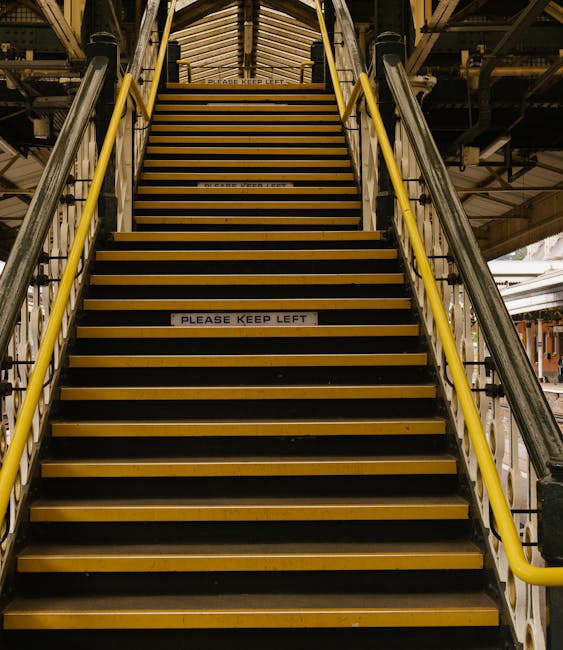 A set of industrial metal stairs with yellow safety railings on either side, leading upwards inside a warehouse or loading area. The stairs have black anti-slip treads with yellow edges and a sign positioned in the middle of the staircase that reads 'PLEASE KEEP LEFT.' The surrounding environment features metal scaffolding and structural elements, with overhead lighting illuminating the staircase. This scene captures a typical access route used during house removals and furniture transport, illustrating the logistic considerations involved in loading goods into a vehicle or navigating a building during a home relocation, with a focus on safety and efficient movement, as part of professional removals services like Westminster Man and Van.